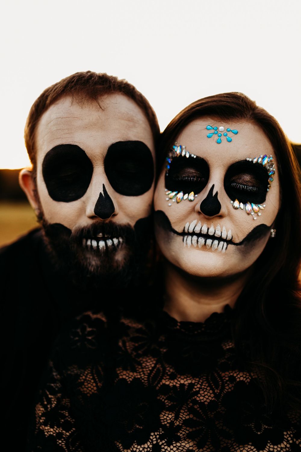 Portrait of couple with skull face paint, woman adorned with blue gems and rhinestones at sunset.