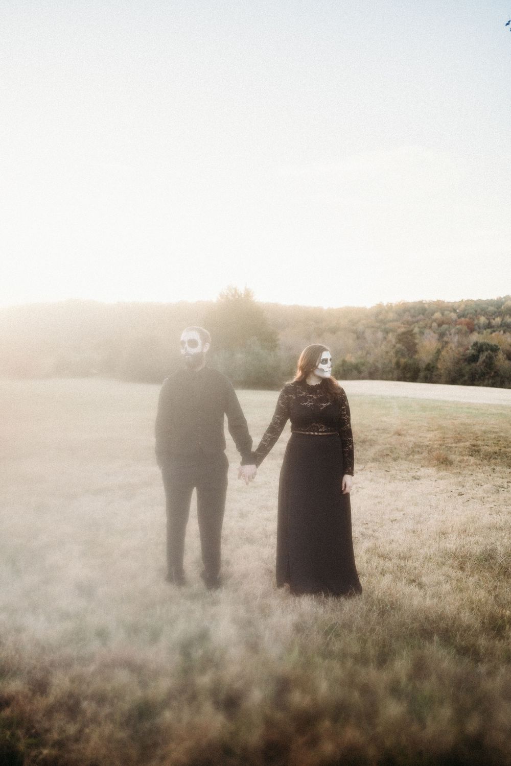 Couple in black outfits and skull face paint hold hands in a misty autumn field.