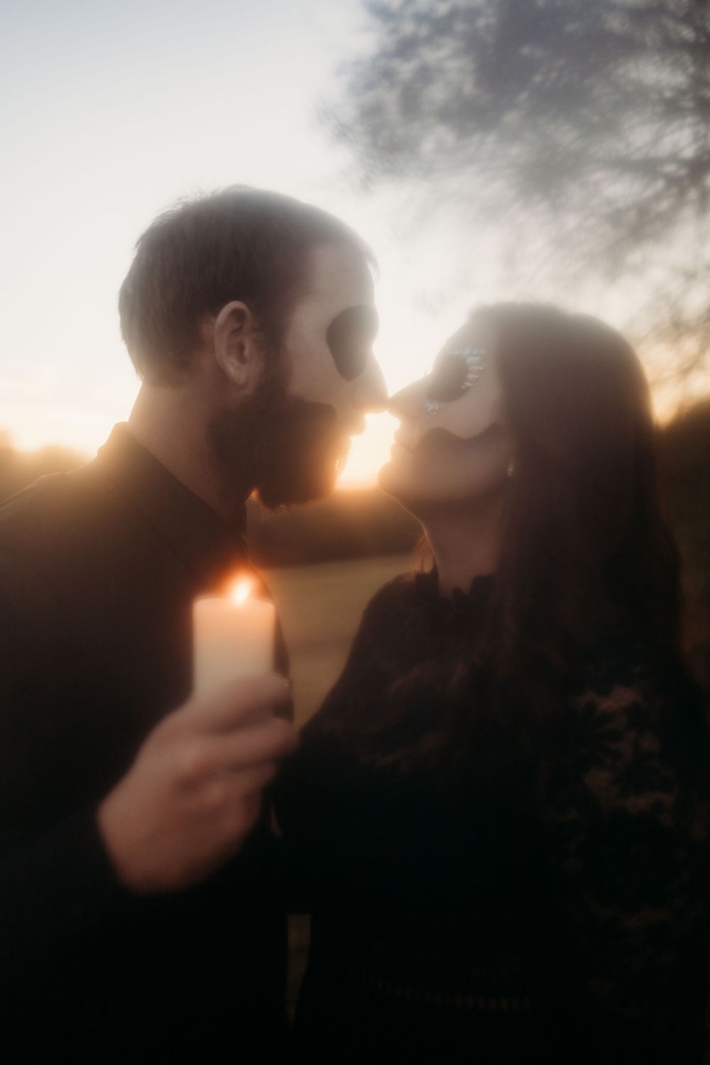 Couple with skull masks share a romantic kiss by candlelight at sunset outdoors.