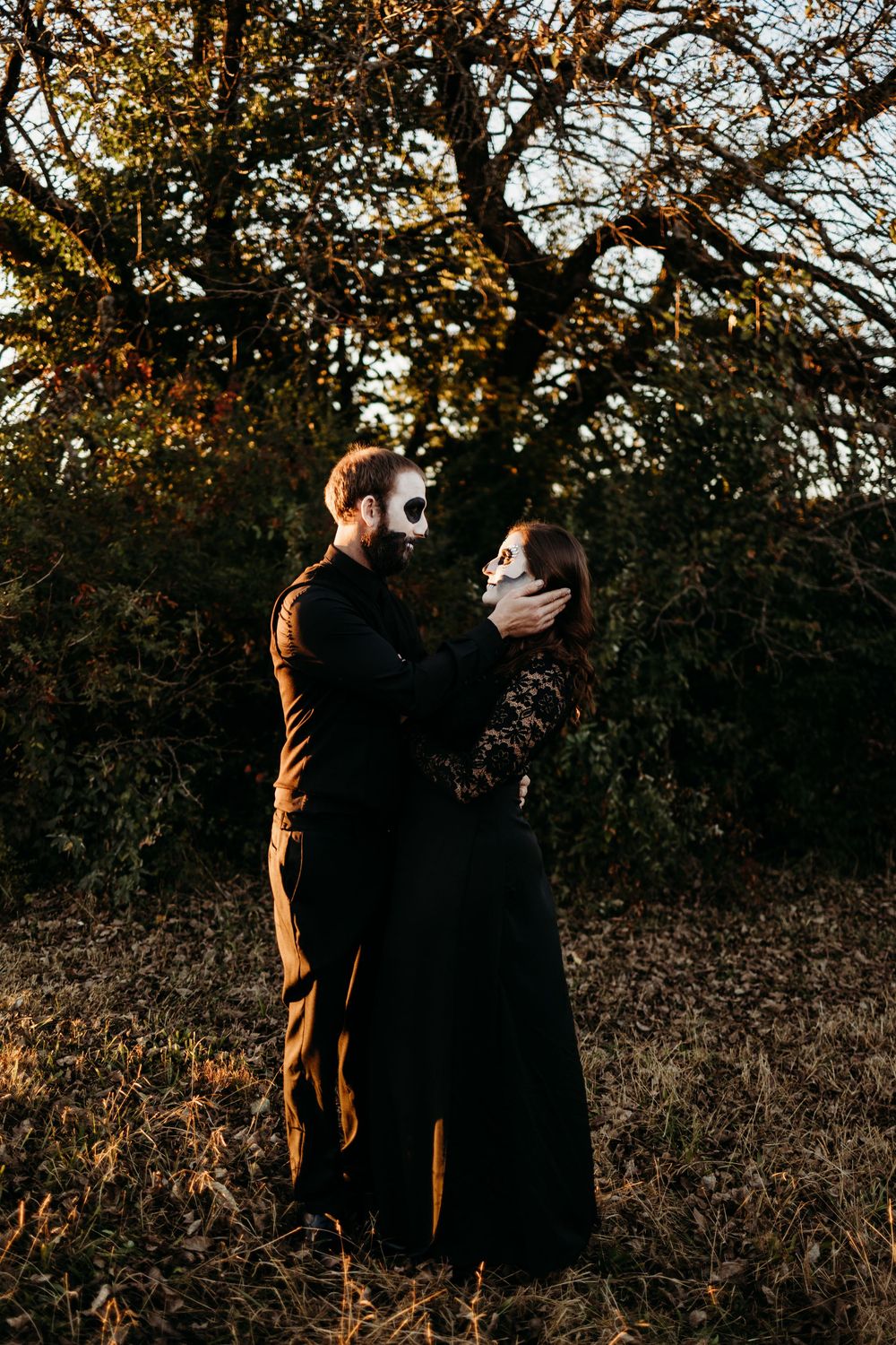 Couple in black outfits and skull face paint embracing in autumn forest at golden hour.