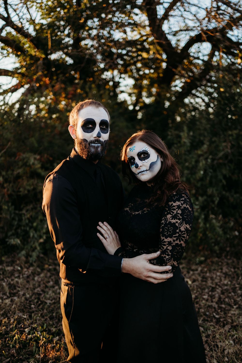Couple wearing black outfits and Day of the Dead skull face paint hugging outdoors in autumn.