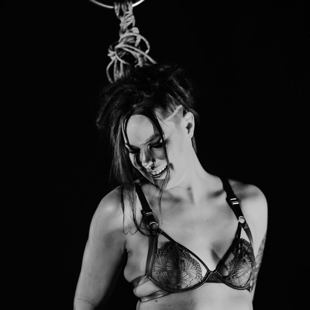 Black and white portrait of a figure wearing a bra and smiling expressively against a dark background.