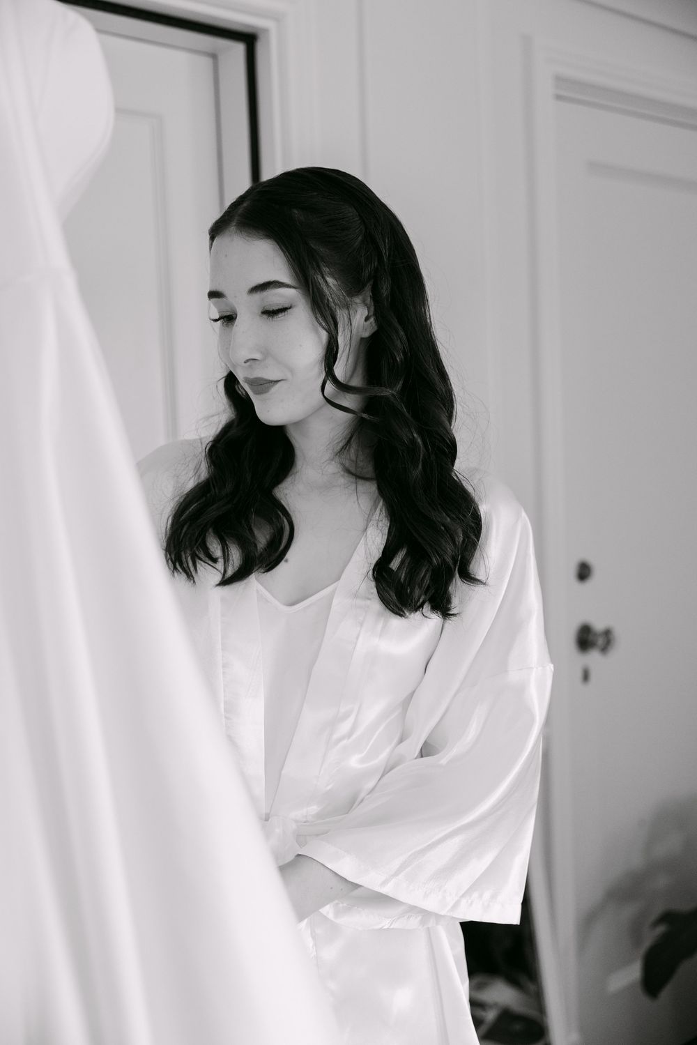 A black and white portrait of a person with long dark wavy hair wearing a white shirt, looking down with closed eyes.