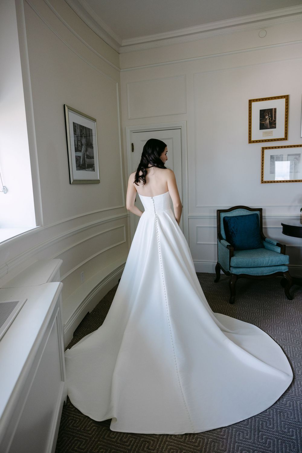 A bride in an elegant white strapless wedding gown with a long train stands in an elegant room with blue armchair.