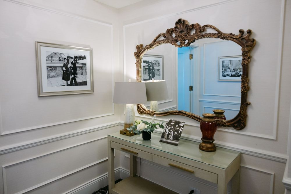 Elegant white dressing room features ornate gold mirror, framed artwork, and classic wainscoting on walls.