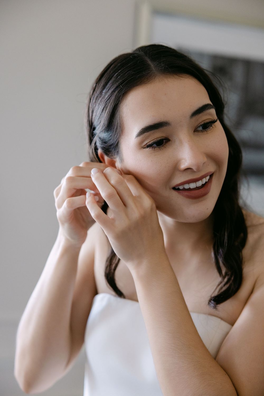 Beautiful bride wearing a white dress smiles while putting on her earrings in a makeup preparation scene.