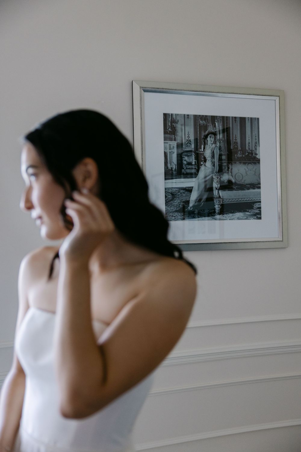 Side view of someone in a white tank top adjusting their earring, with a framed black and white photograph on the wall.