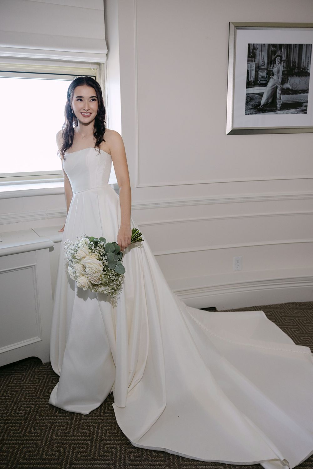 A person in an elegant white wedding gown with a long train holds a bouquet of white flowers beside a window.