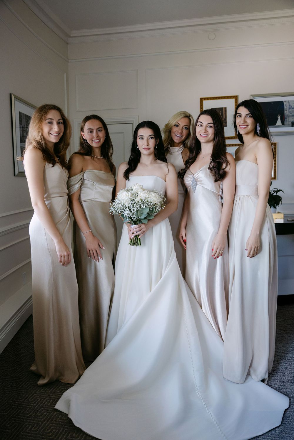 A bridal party in neutral-toned dresses poses together in an interior setting with a floral bouquet.