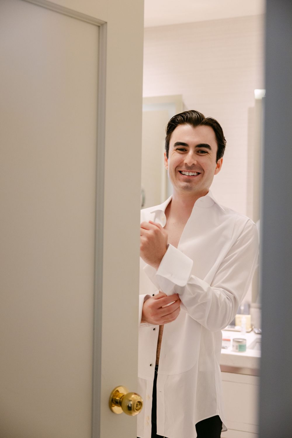 Smiling person in a crisp white dress shirt stands in a doorway while getting dressed.