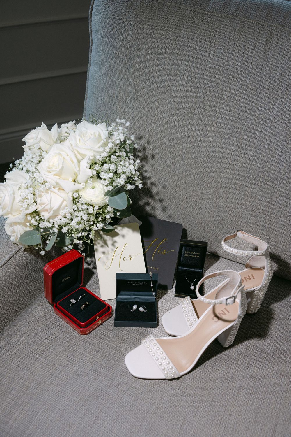 White bridal shoe, floral bouquet, and wedding rings in jewelry boxes displayed on gray fabric backdrop.