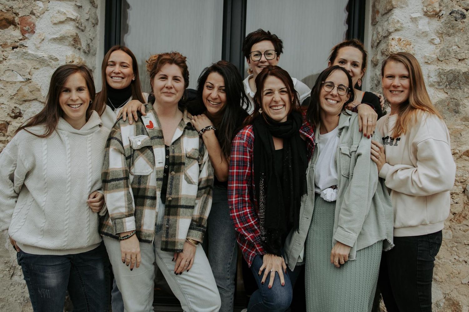A group of people wearing casual winter clothing stand together laughing and smiling against a stone wall.