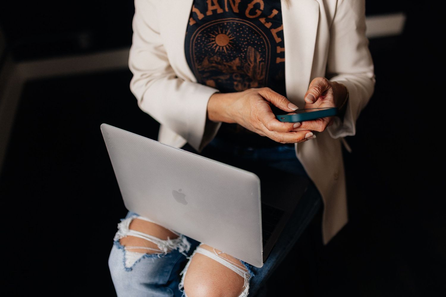 Person in ripped jeans works on laptop while holding smartphone, wearing casual business attire.