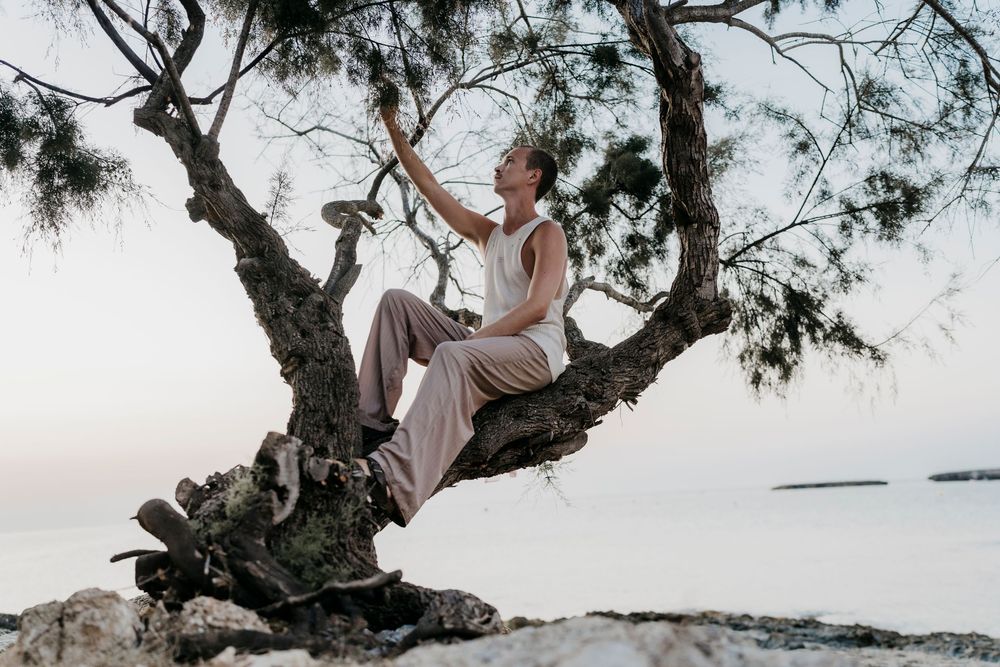Person performing expressive dance movements in pink clothing among coastal trees.
