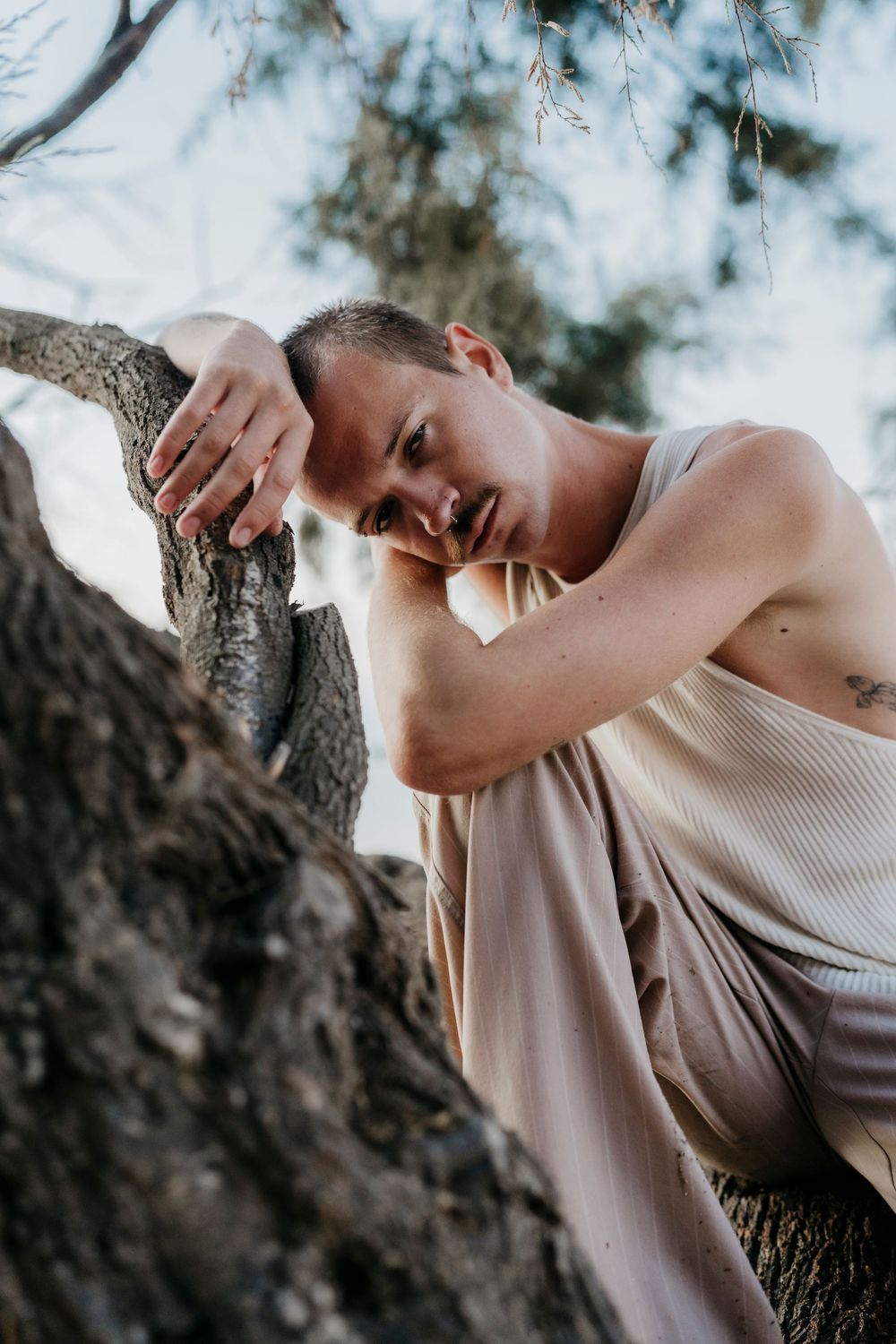 Person in beige tank top leaning artistically against tree trunk near ocean.