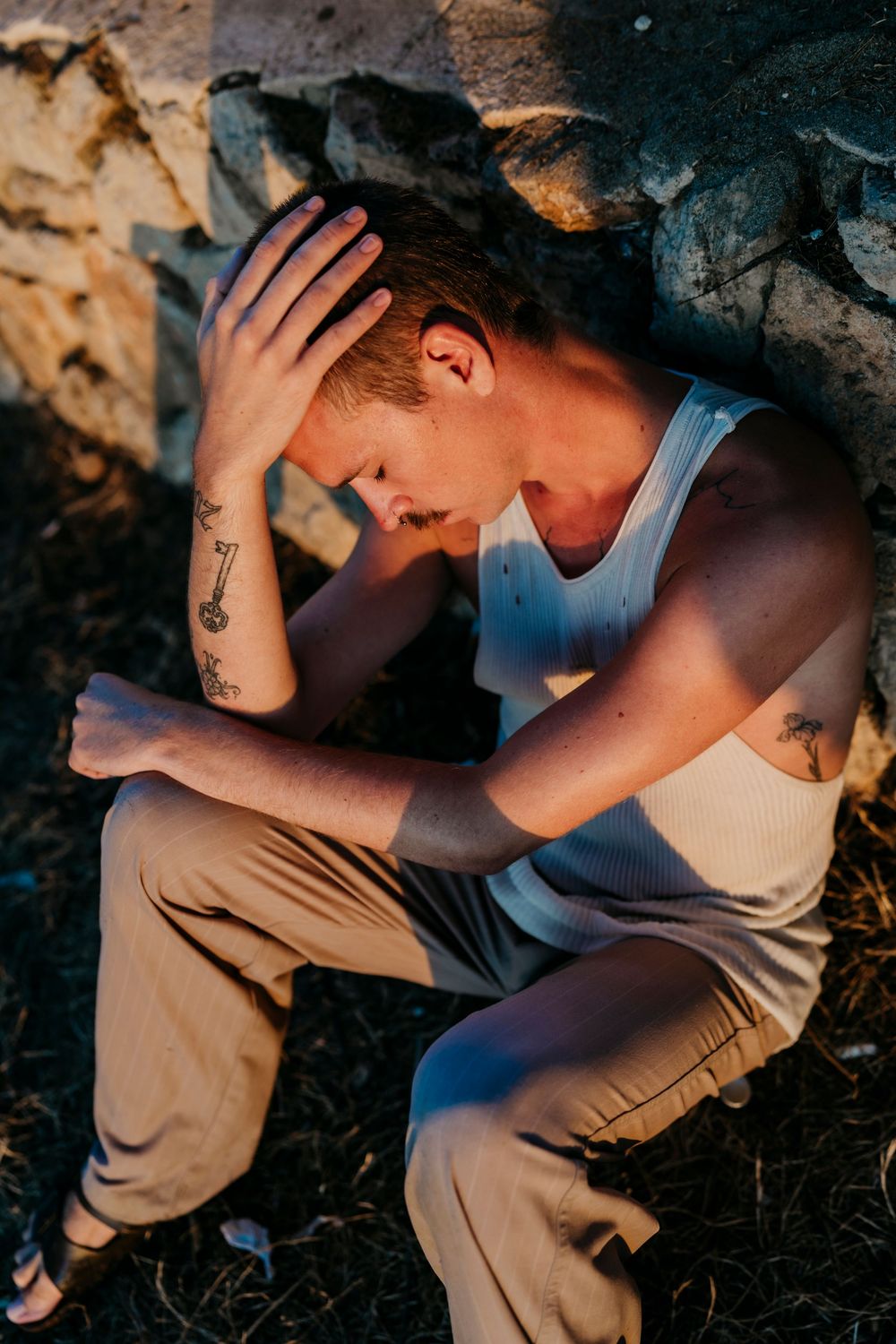 Person in white tank top sitting against stone wall with hand on head.