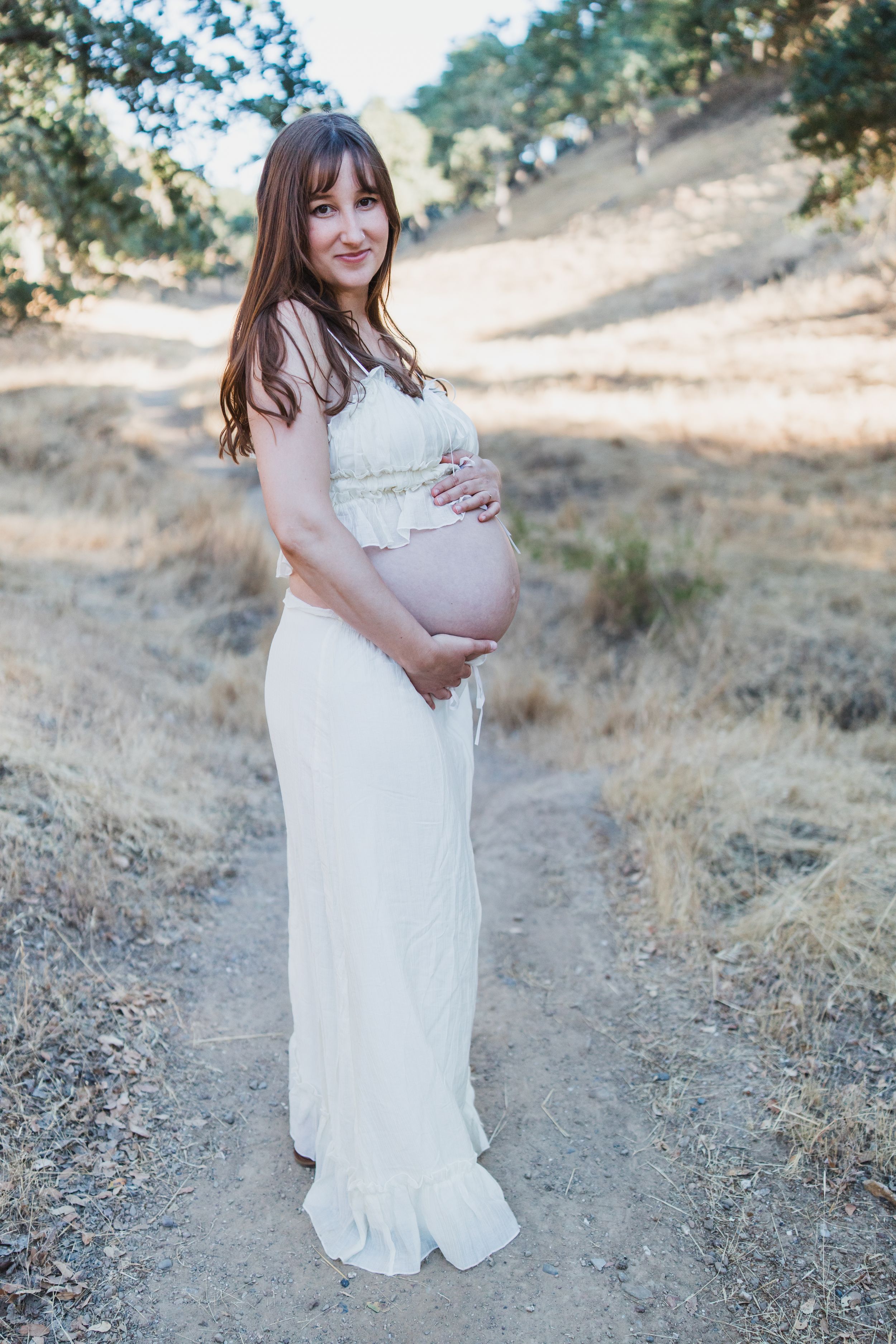 Expectant mother wearing a flowing white lace maternity gown poses on a dirt path surrounded by golden hills.
