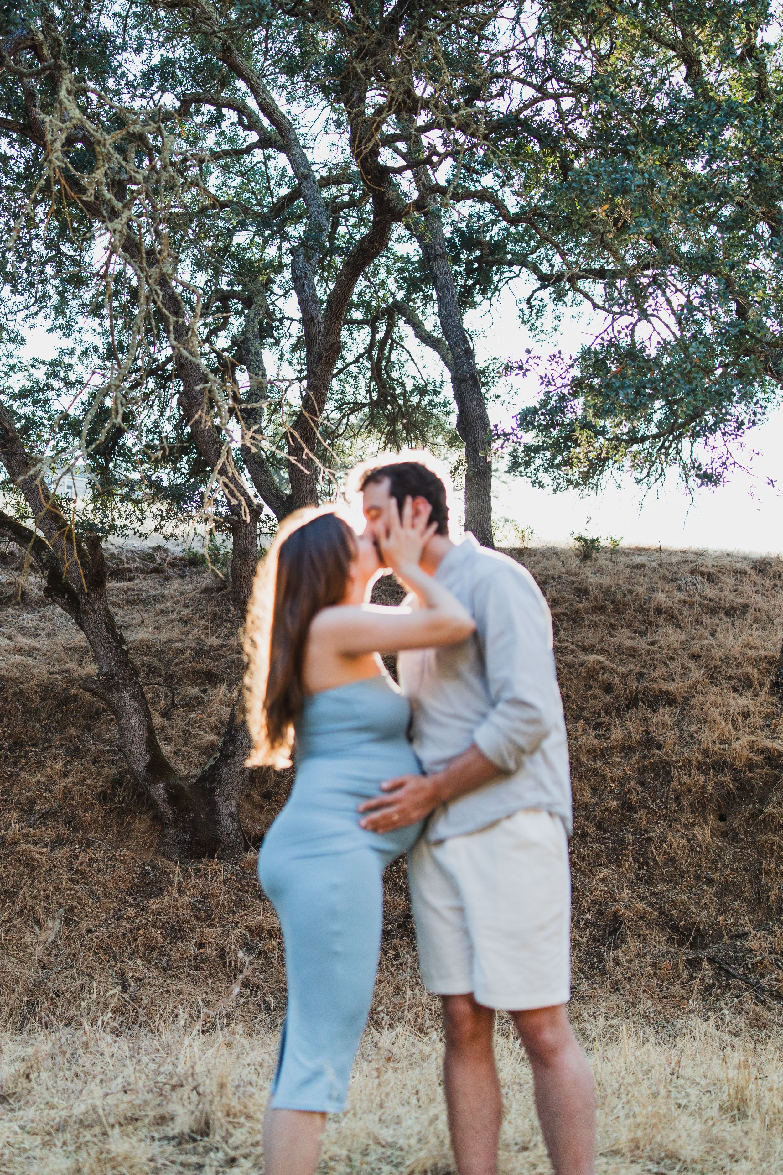 Romantic couple in a blue dress and beige outfit share tender moments under trees at sunset in a natural setting.