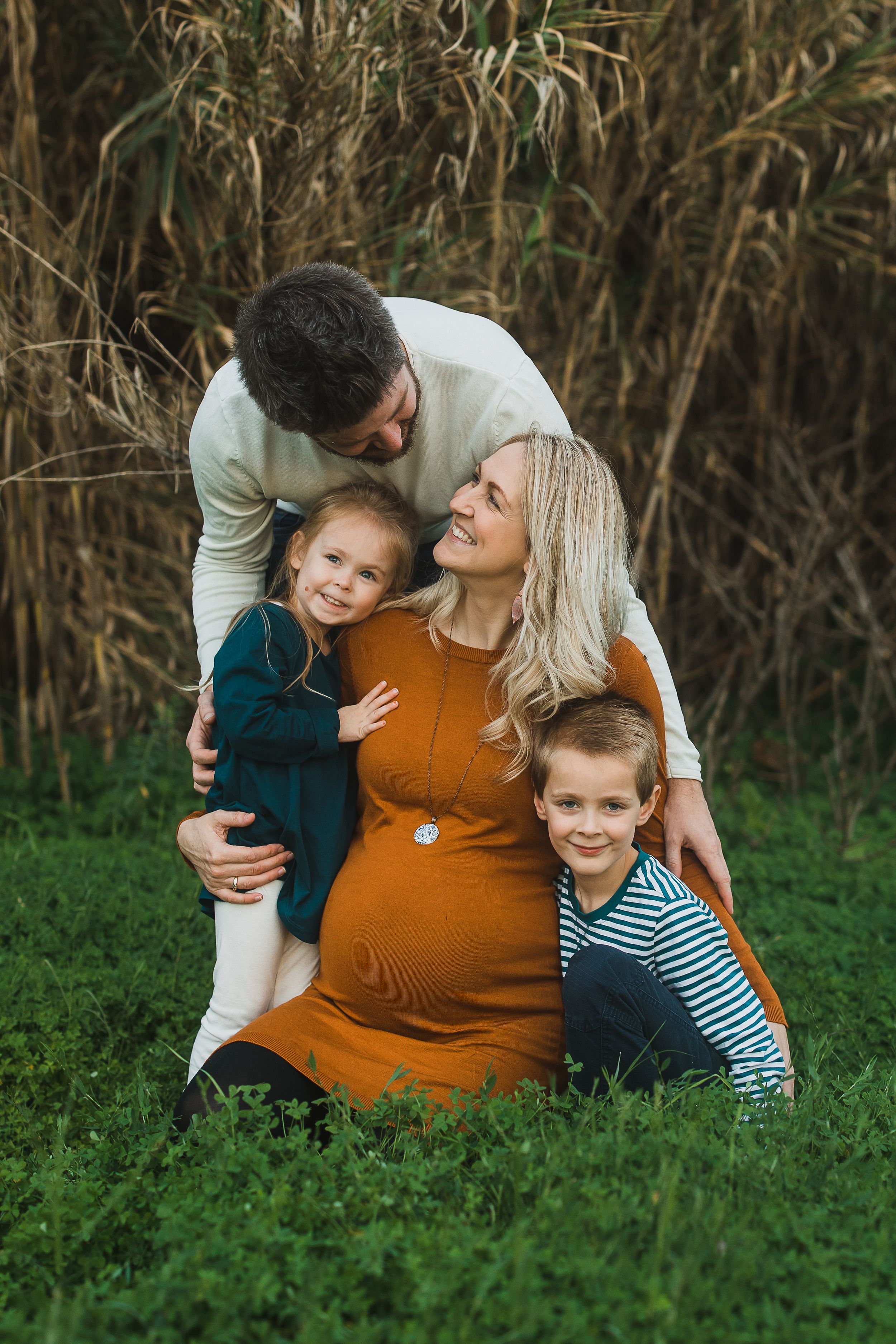 Family embraces while sitting together in a grassy field with golden light filtering through brush in the background.