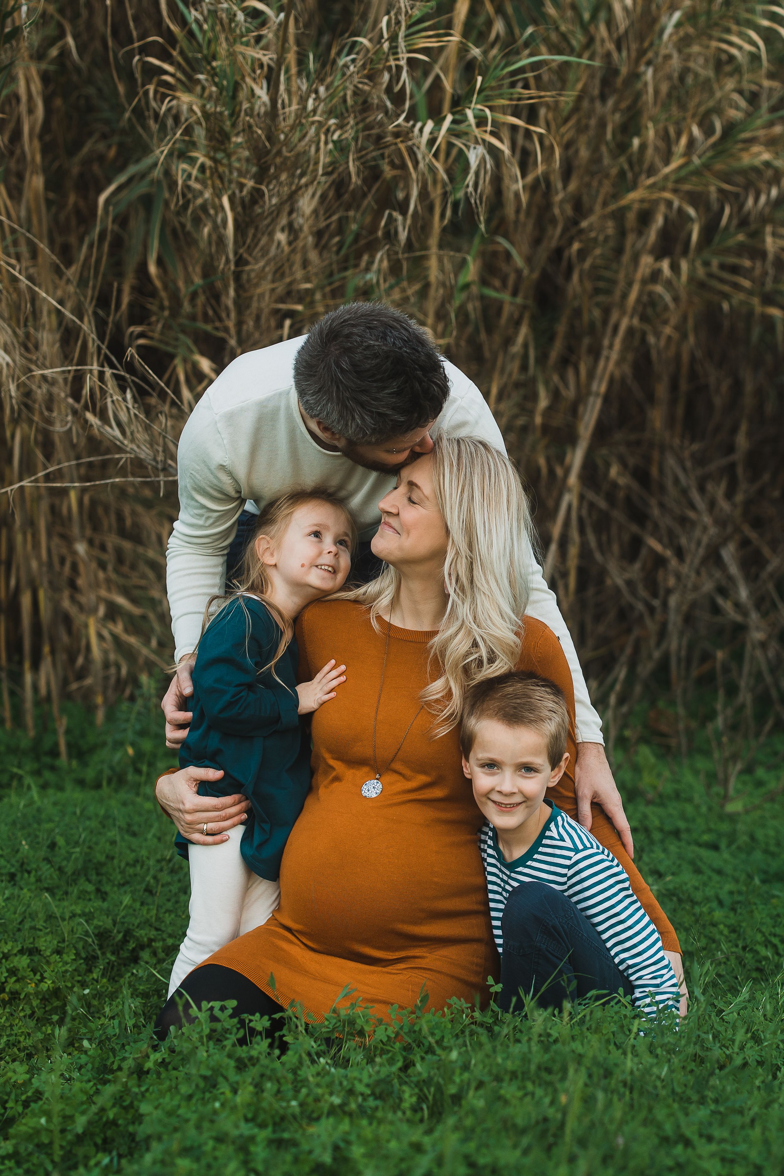 Family shares tender moments as they lay together on green grass with tall reeds in the background.