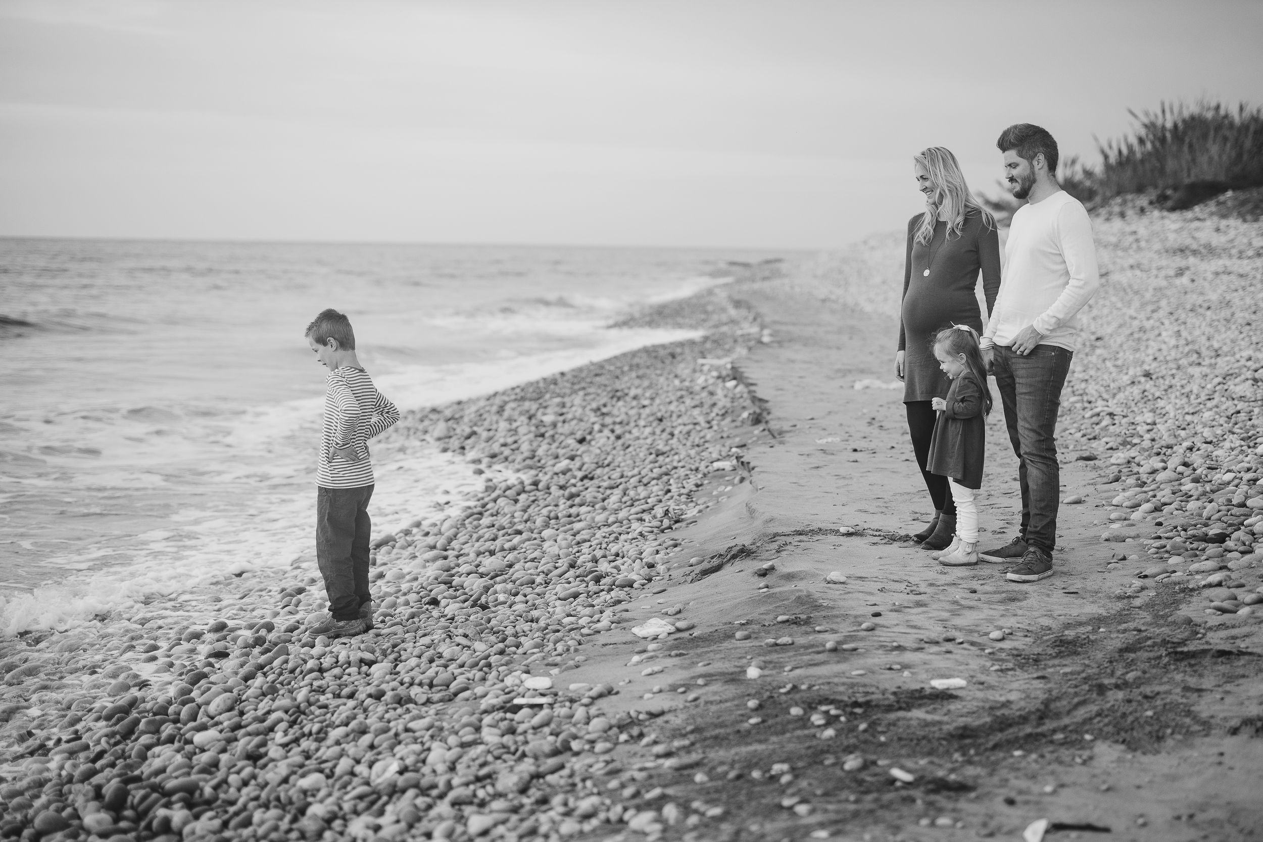 People walking along a scenic beach shoreline during sunset with crashing waves and pebbles in black and white.