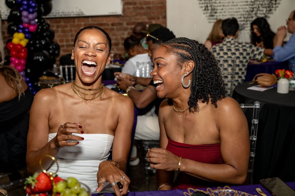Two friends laughing joyfully at a festive party with colorful decorations in the background.