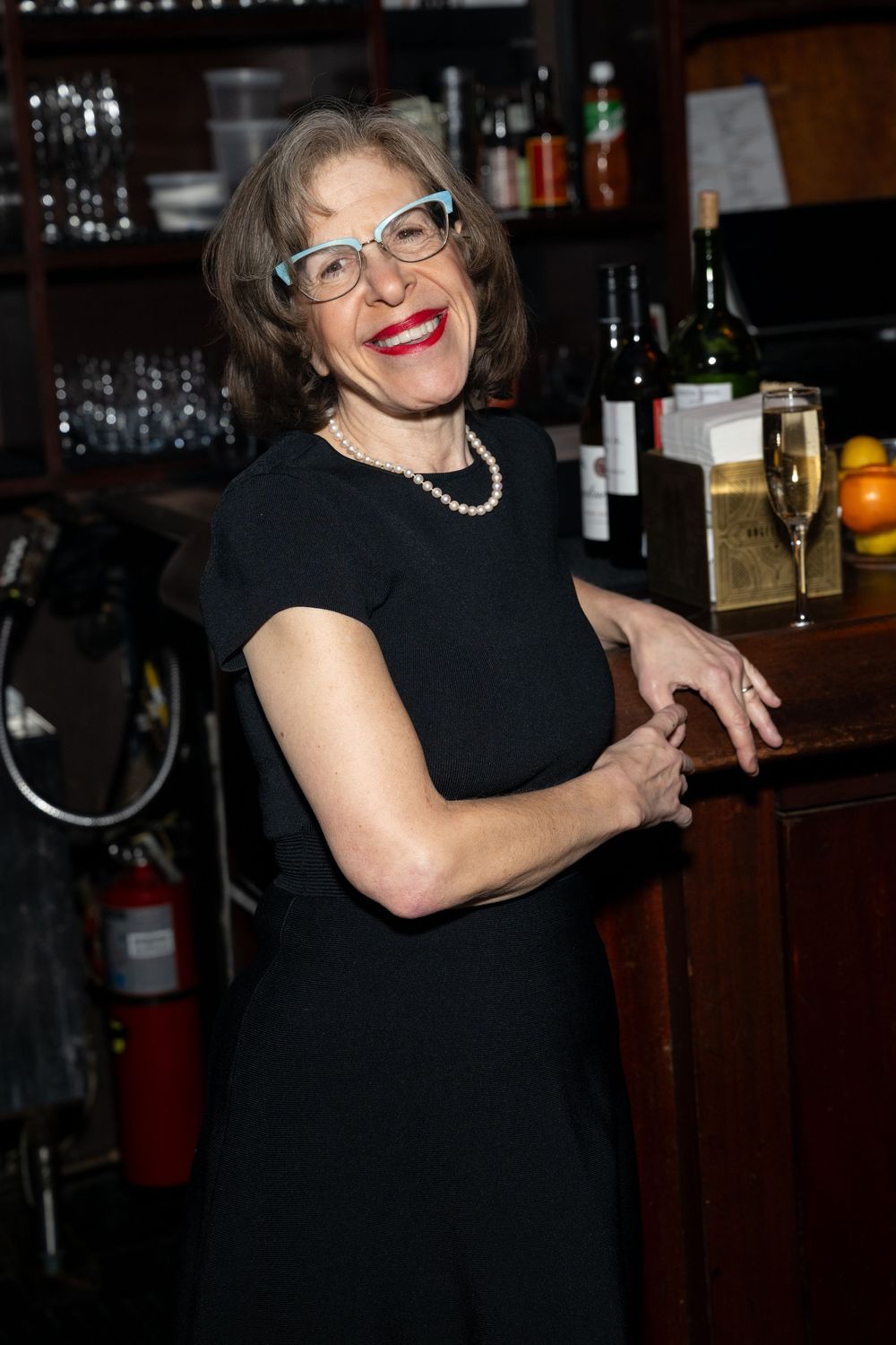 A person in a black dress and glasses poses playfully at a bar in a series of candid shots.