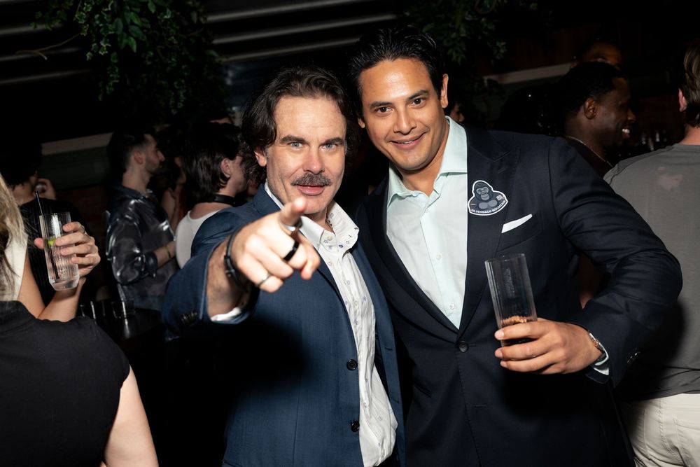 Two men smiling and posing at an indoor party event, one pointing at camera while holding drinks.