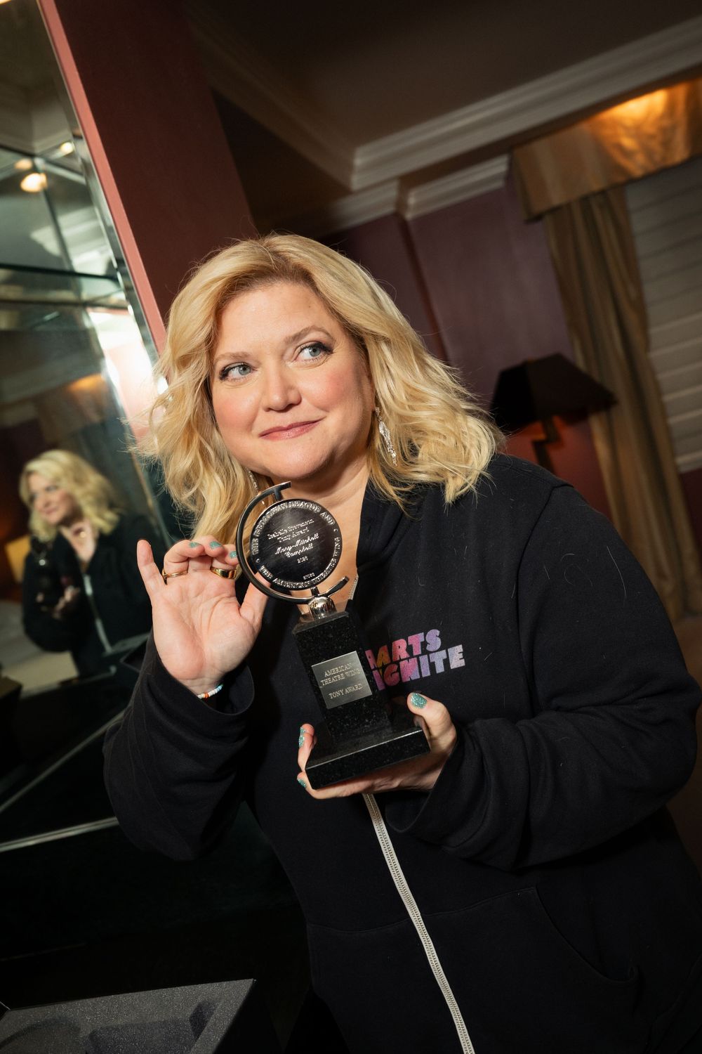 Woman in Arts Ignite hoodie proudly holding a circular award trophy, smiling at an indoor event.