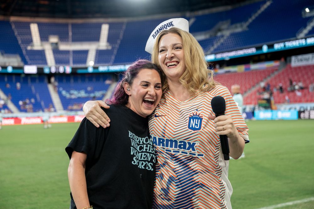 Two women smiling and posing together on a soccer field, one holding a microphone wearing an NY jersey.