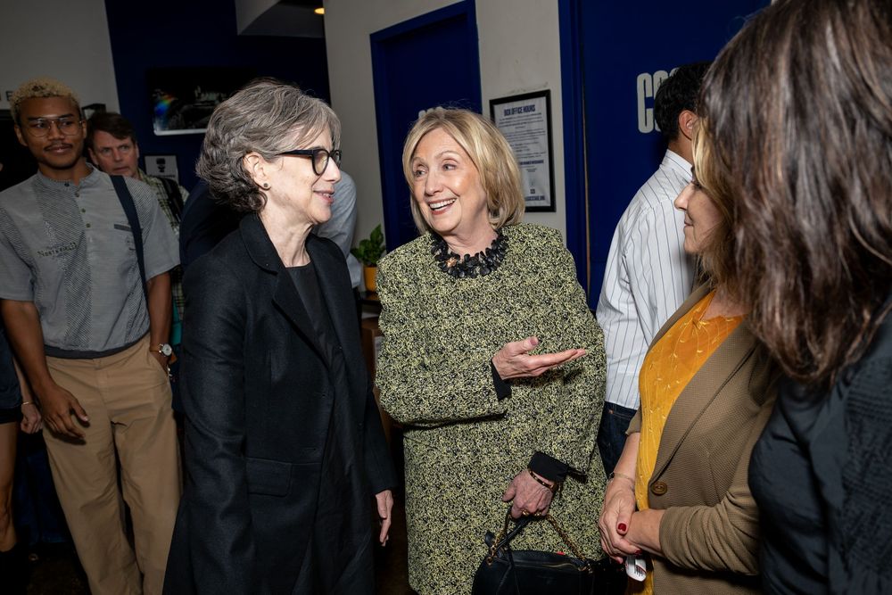 Three women engaged in conversation at an indoor networking event with people gathered in the background.