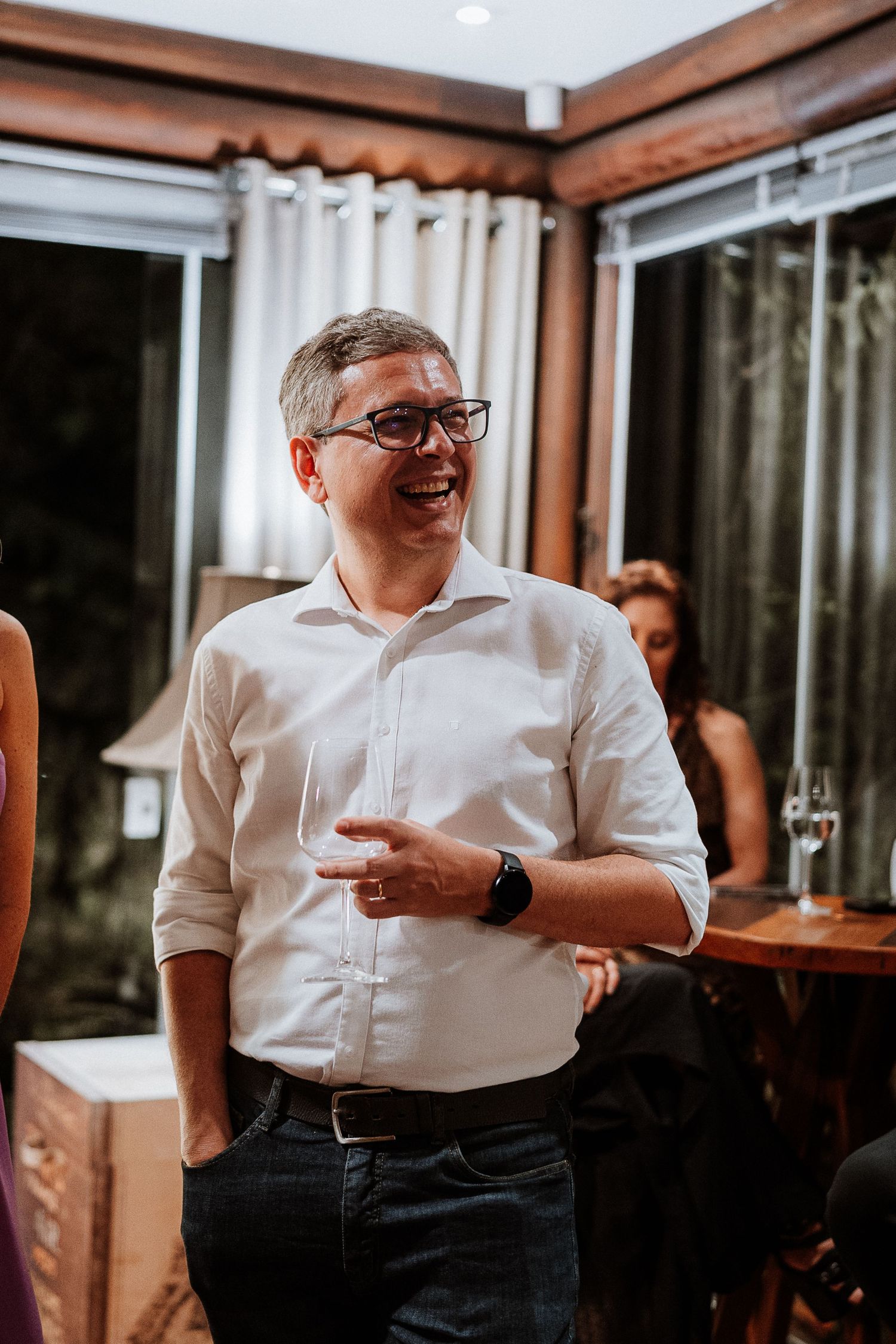 Person in white shirt and glasses gesturing during indoor gathering.
