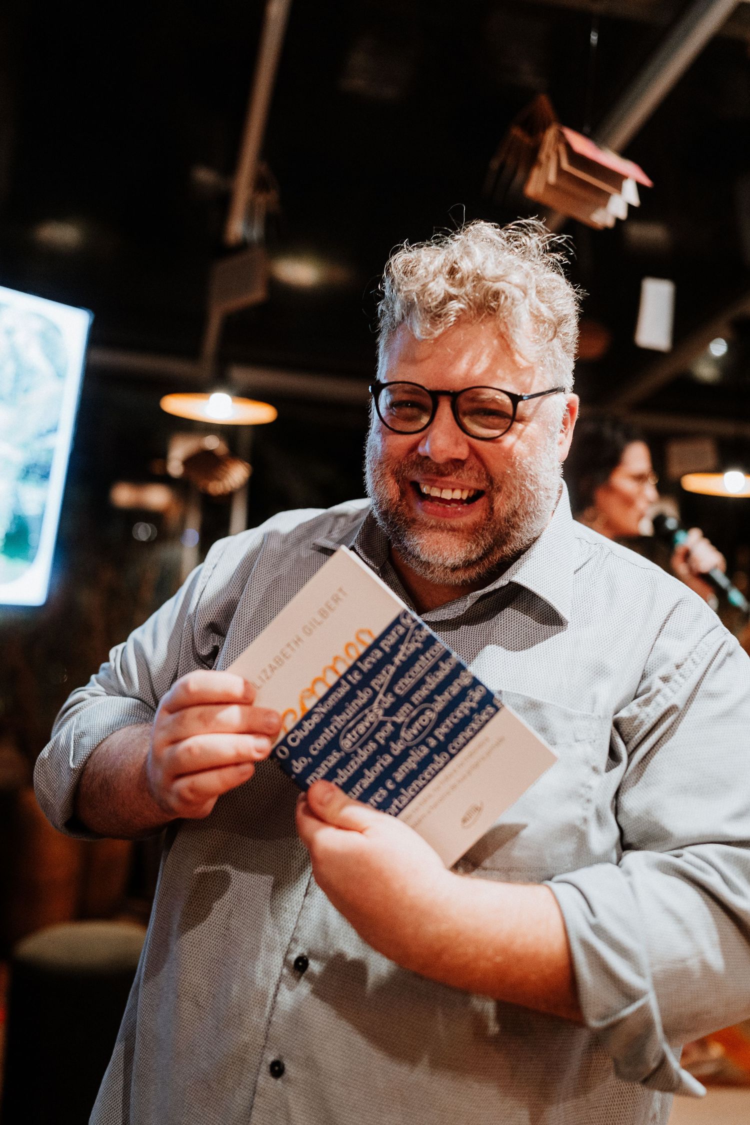 Person in glasses holding a book and smiling warmly at an indoor gathering.
