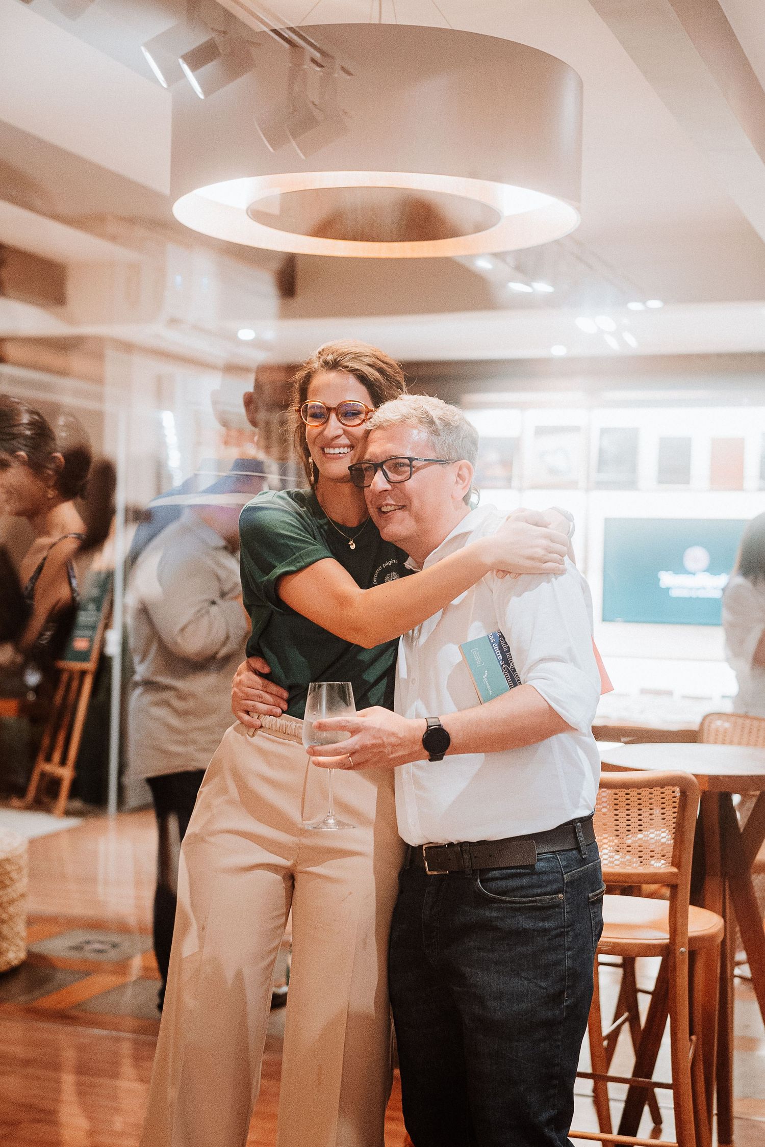 Two people share a warm embrace in a modern office space with circular lighting fixture overhead.