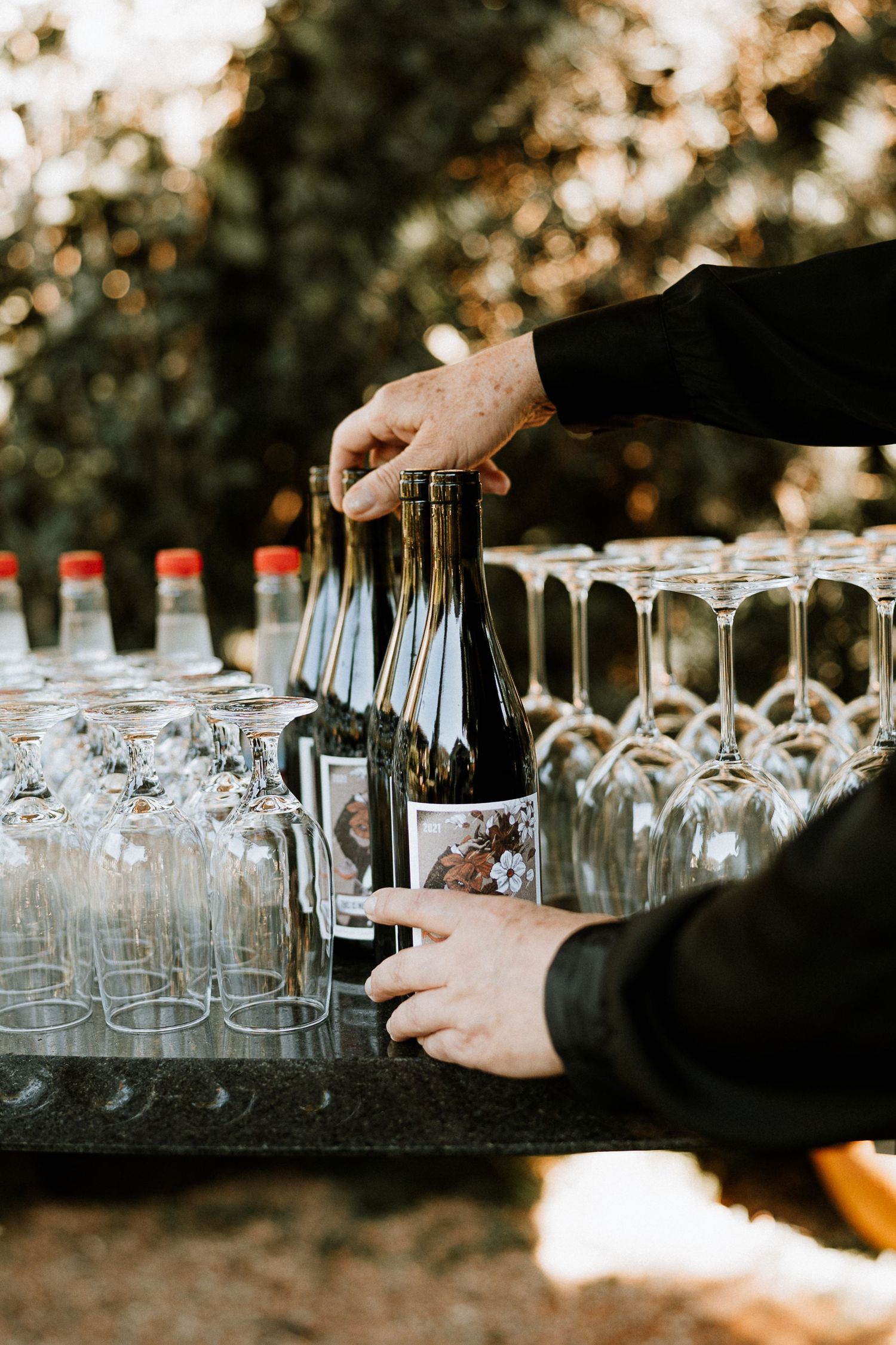 Hands pouring wine bottles at an outdoor event with wine glasses arranged on a table.