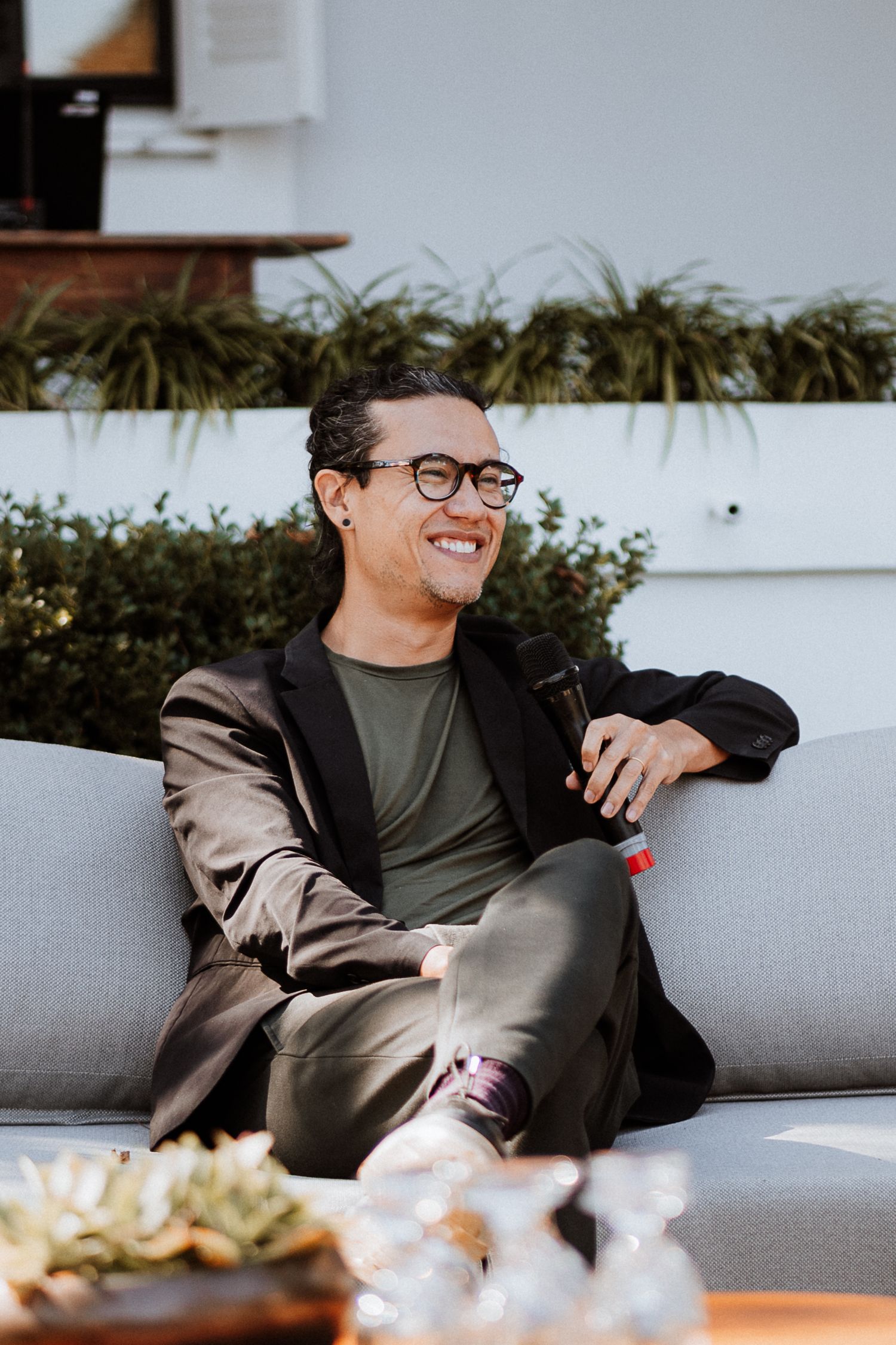 A person in glasses and blazer smiles while relaxing on a gray outdoor couch.