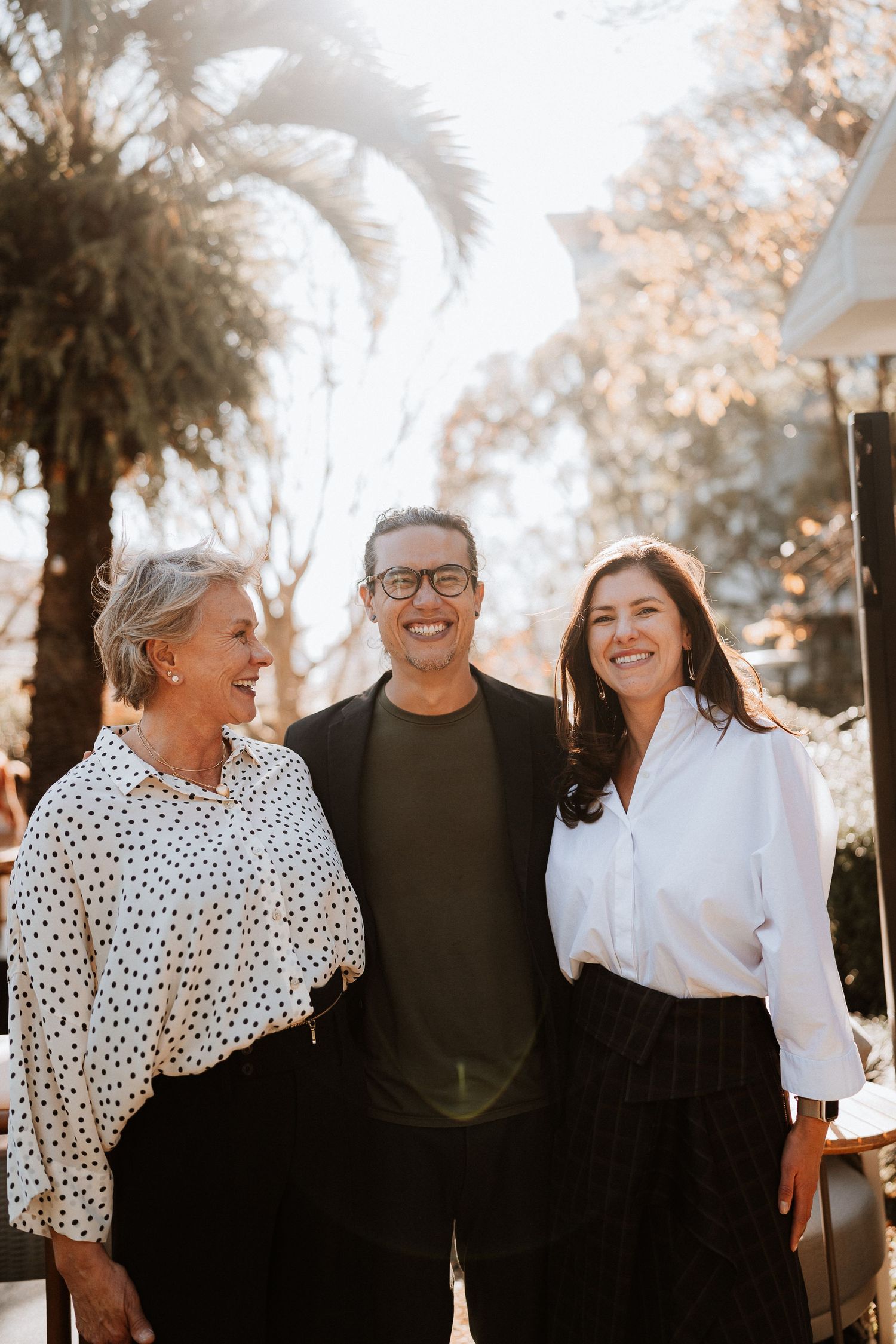 Three people share a joyful moment outdoors with palm trees in background.