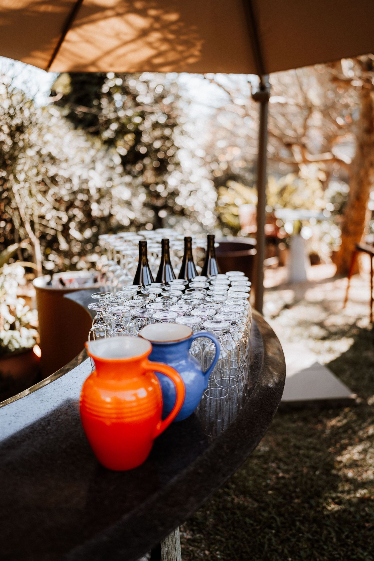 Glass bottles and an orange pitcher arranged on a dark surface under dappled sunlight.