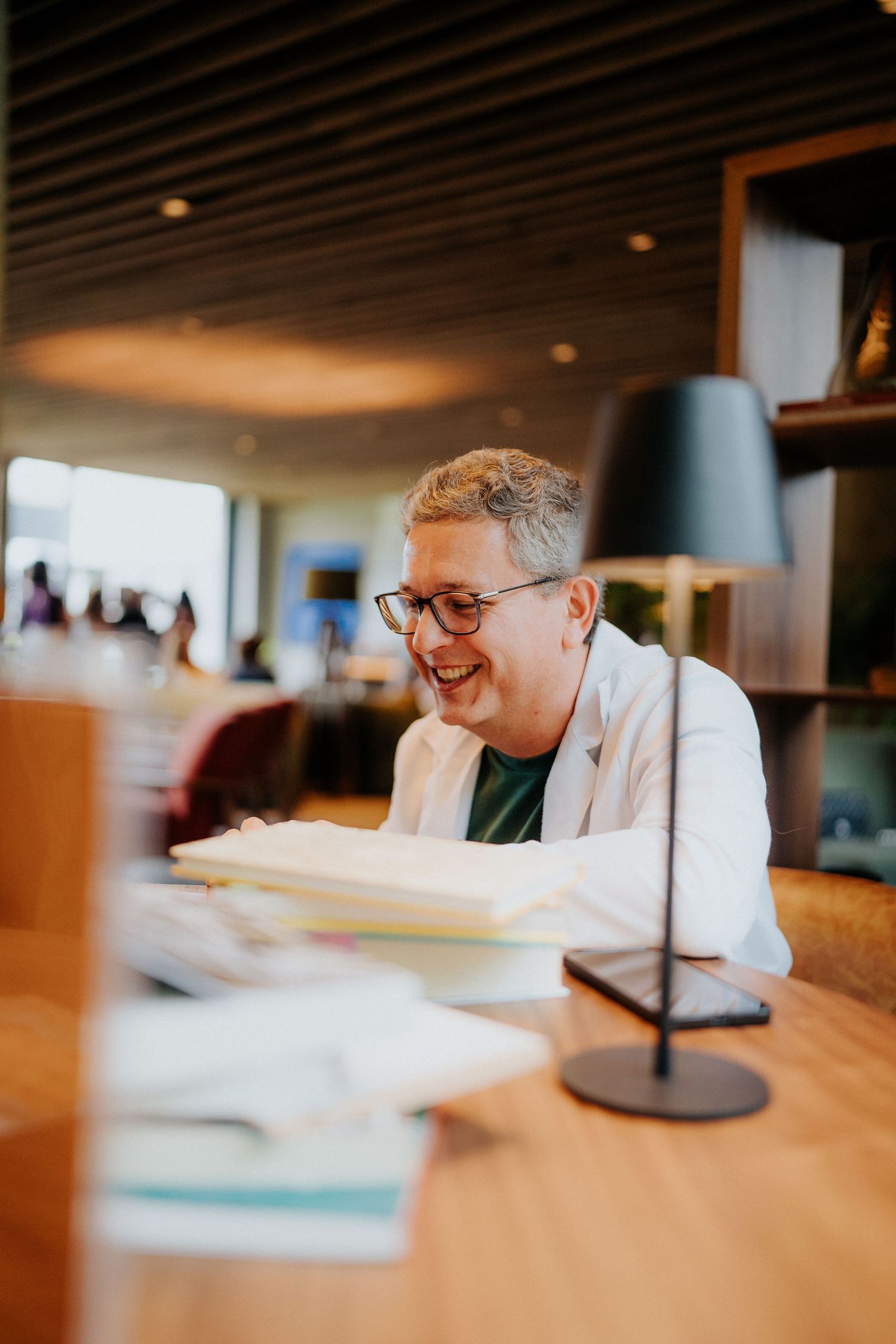Person in white blazer and glasses smiling while seated at a table in a warmly lit restaurant interior.