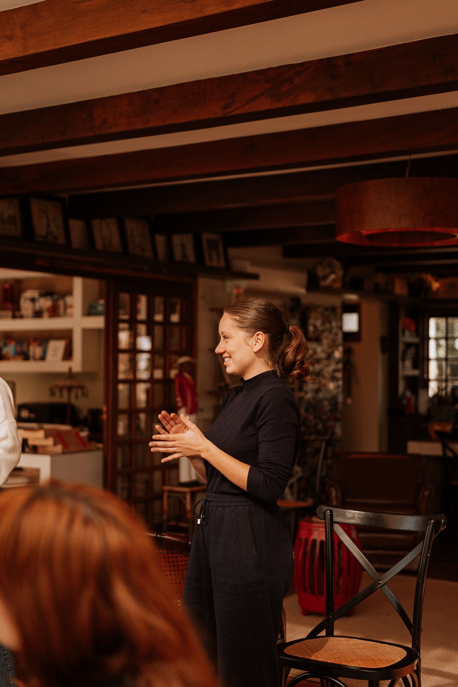 Person in black attire gesturing while speaking in rustic interior setting.
