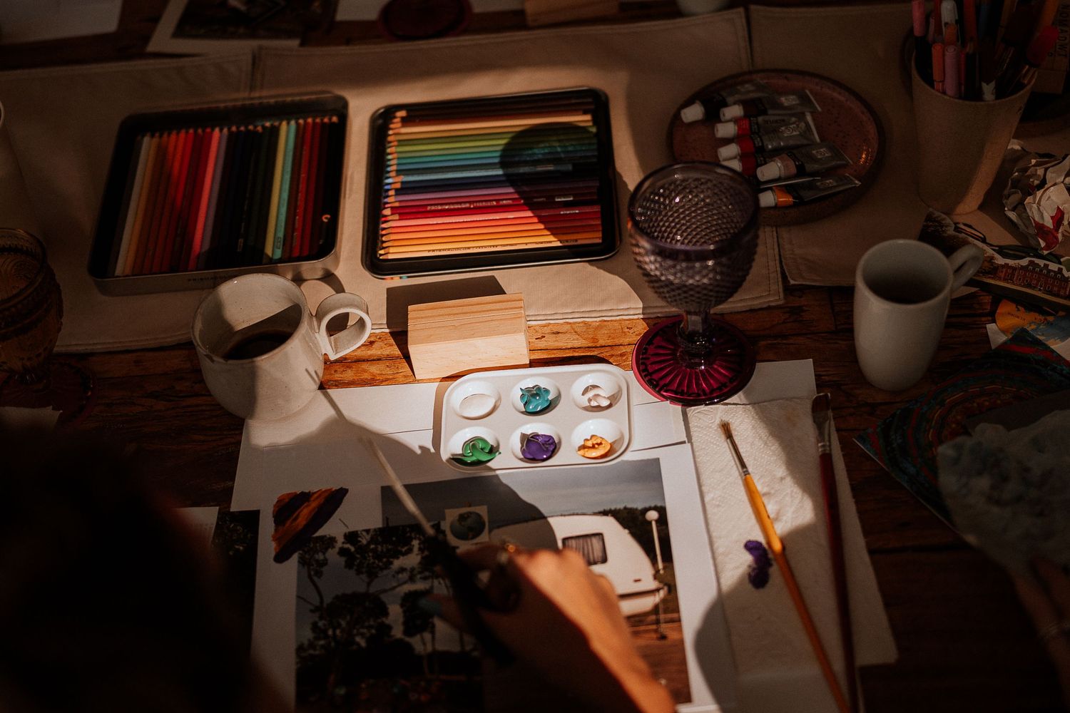 Art supplies and colored pencils arranged on a wooden desk with notebooks and a glass.