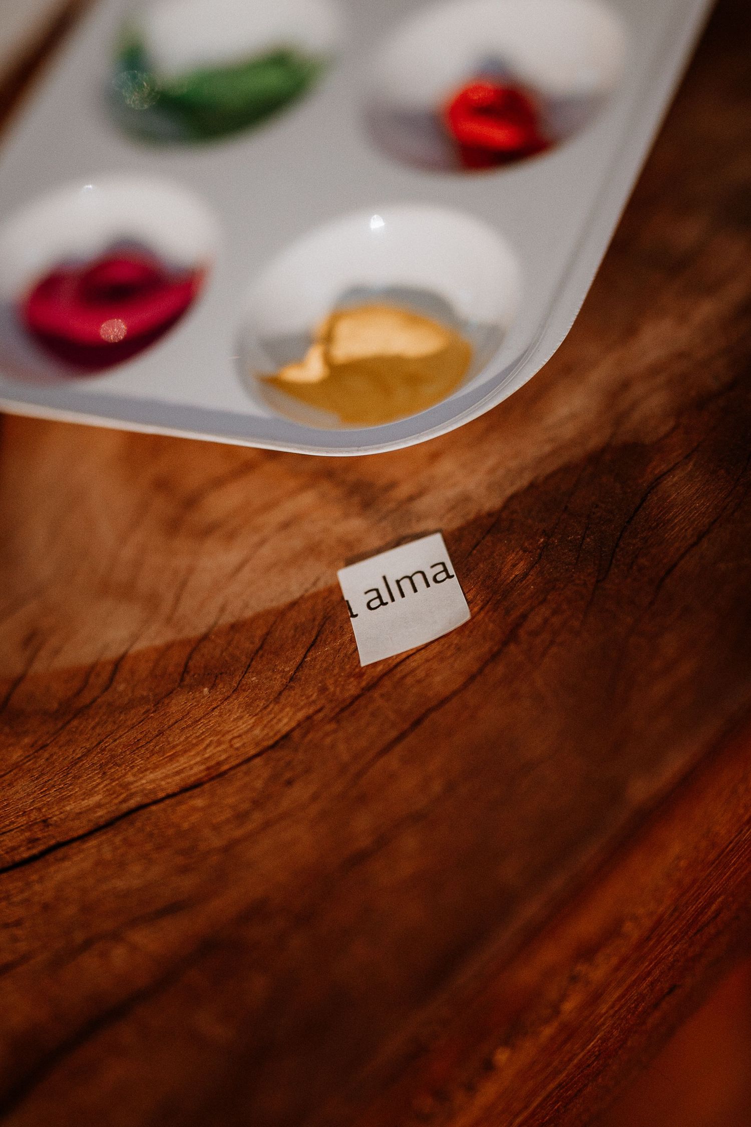 Close-up of colorful paint or pigments in a white palette tray with 'alma' text on wooden surface.