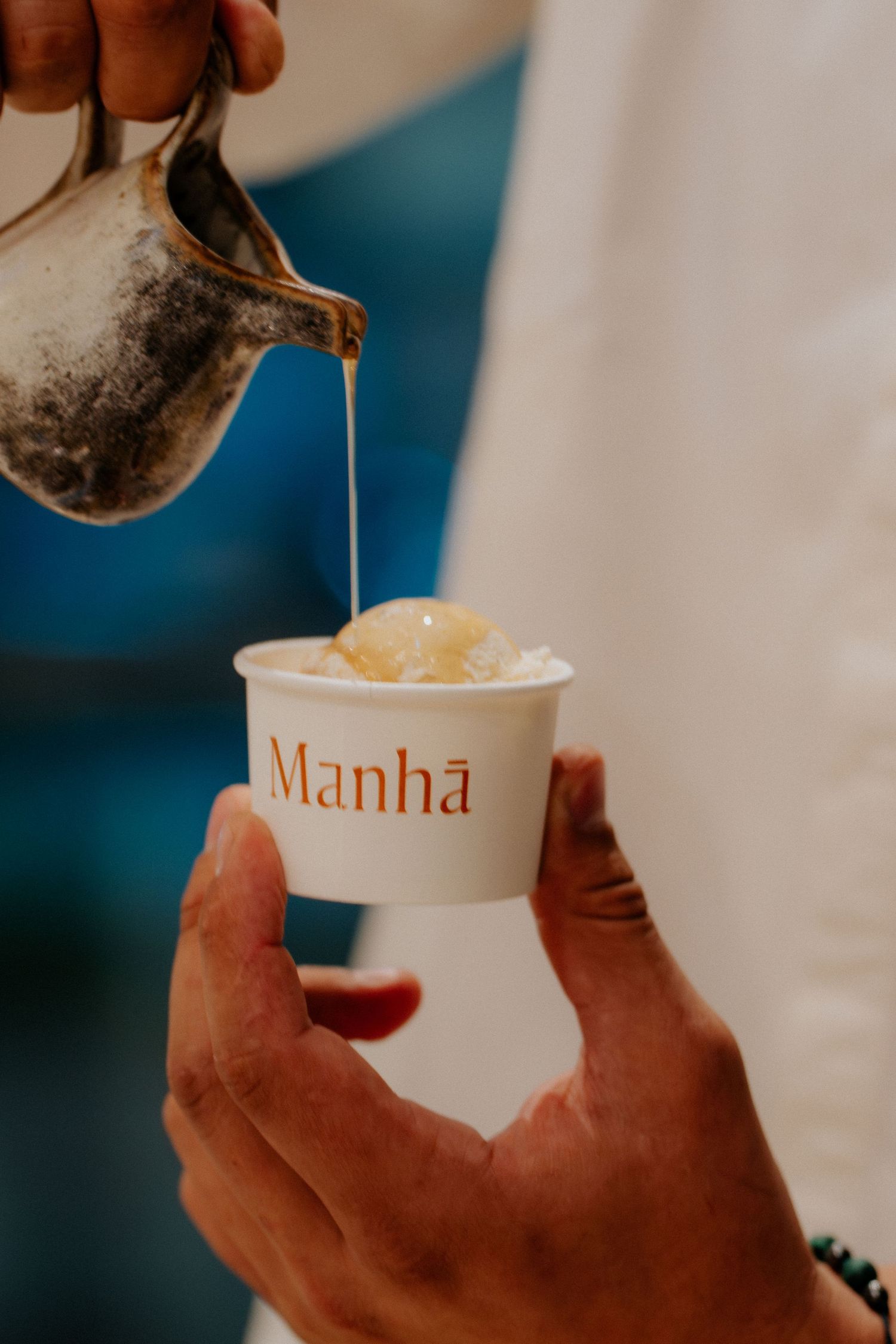 A metallic pitcher pouring liquid into a white cup labeled Manha.