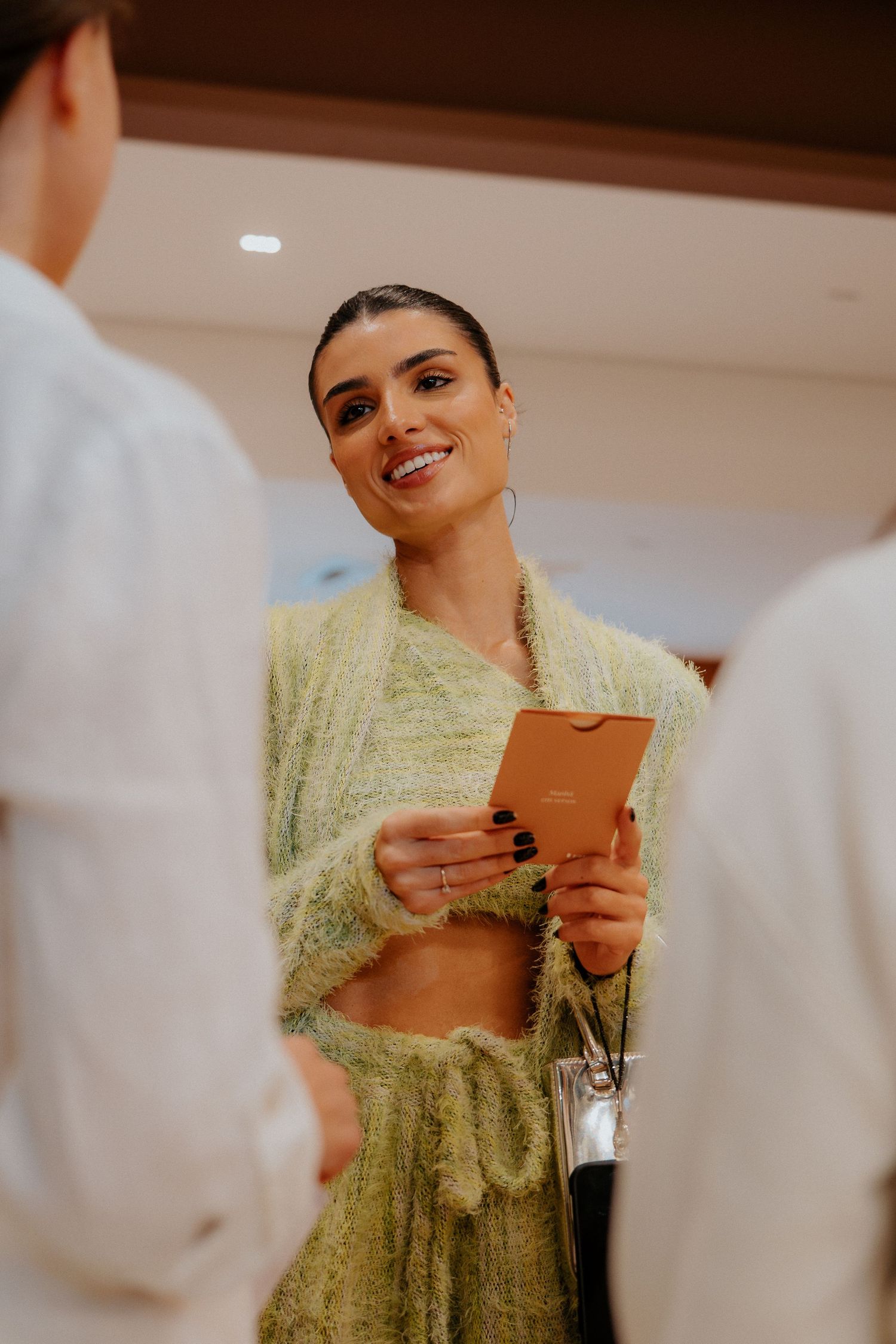 A person in a pale green dress smiles while examining a small card at an event.