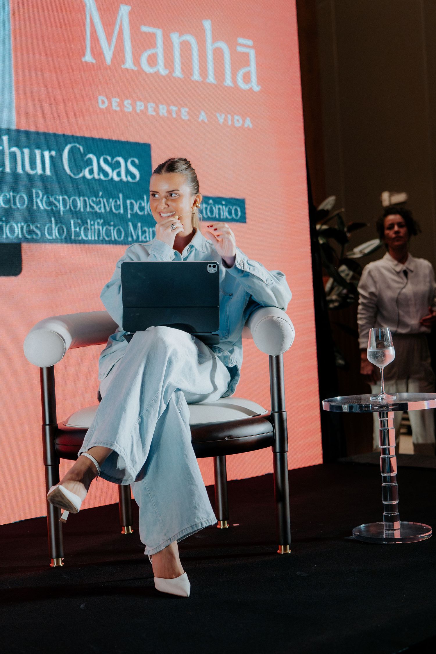Professional speaker seated in white chair at business conference with coral-colored backdrop.