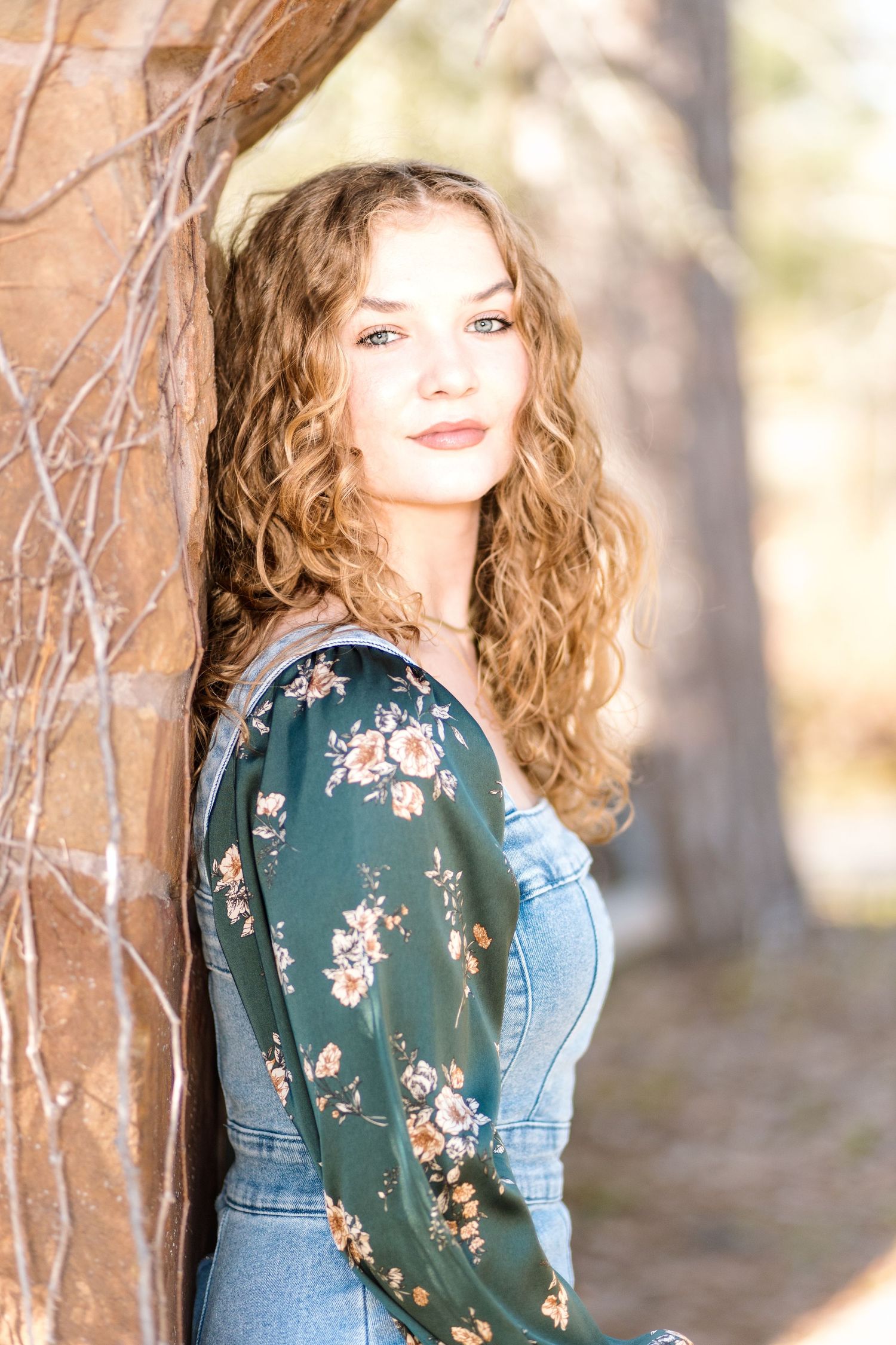 Portrait of a person with wavy hair wearing denim overalls and a green floral blouse in a soft outdoor setting.