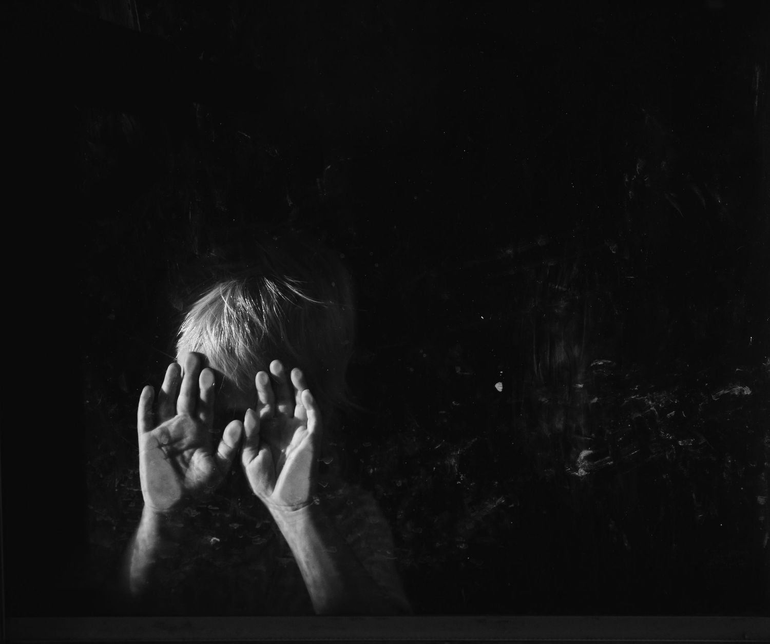 Photo of a toddler hiding behind hands against window in mysterious light in an everyday backyard in Tulsa, Oklahoma