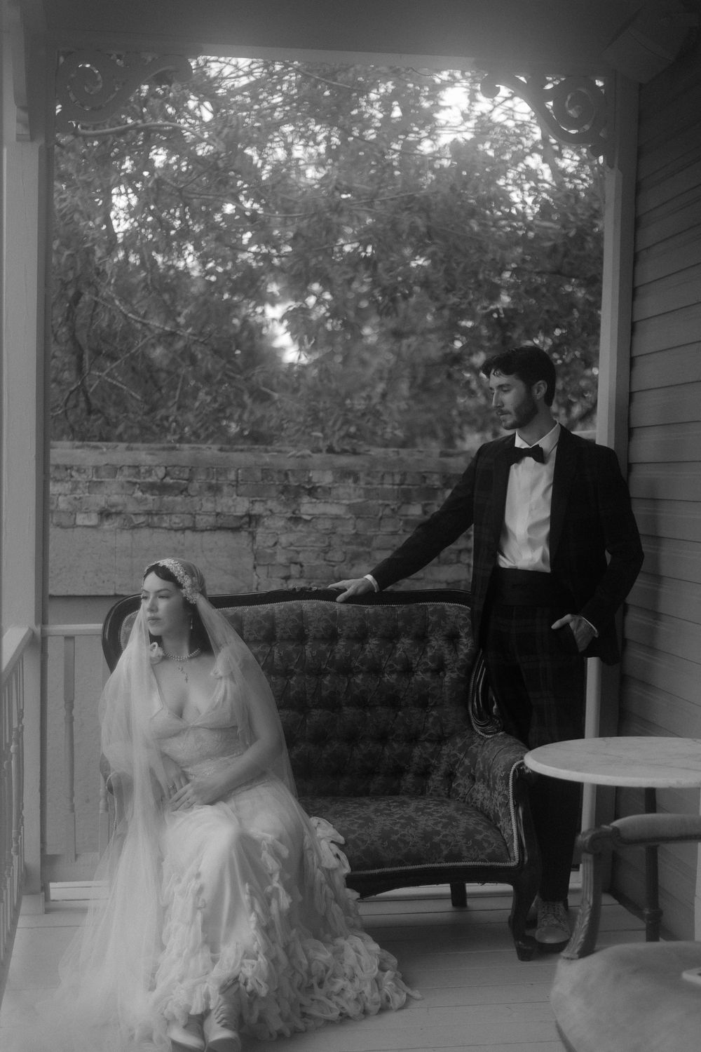 A black and white photo showing a bride and groom in formal attire on an elegant porch with vintage furniture.