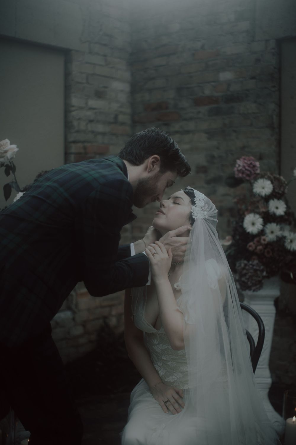 A romantic wedding couple shares a tender kiss in a dimly lit room with exposed brick walls and floral decor.