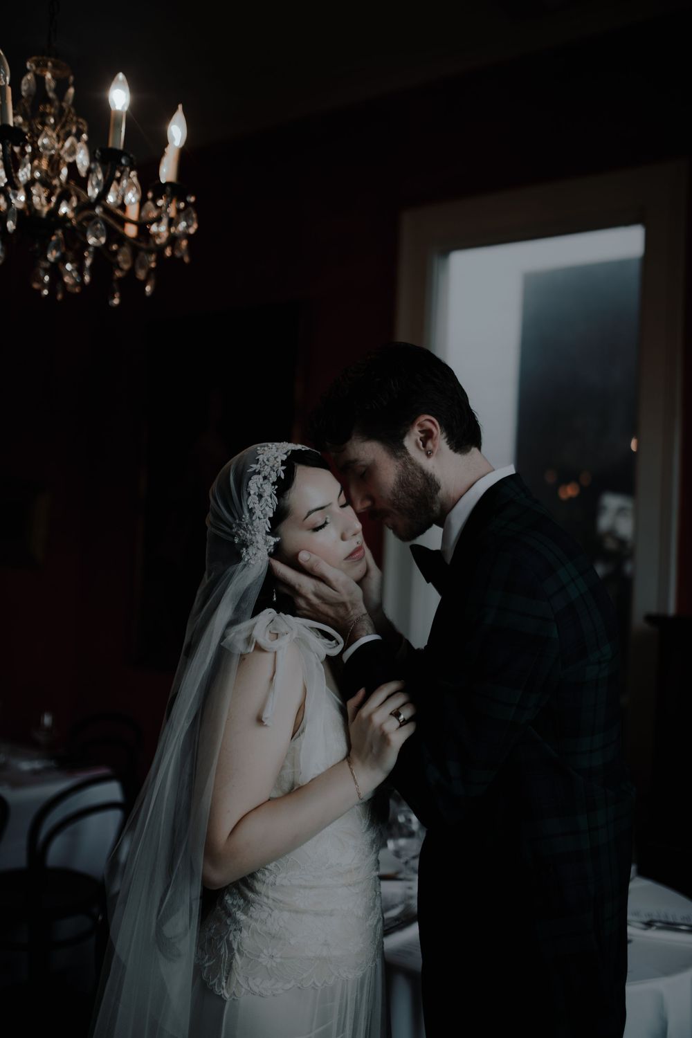 Romantic wedding couple embracing under chandelier in dark, atmospheric setting.